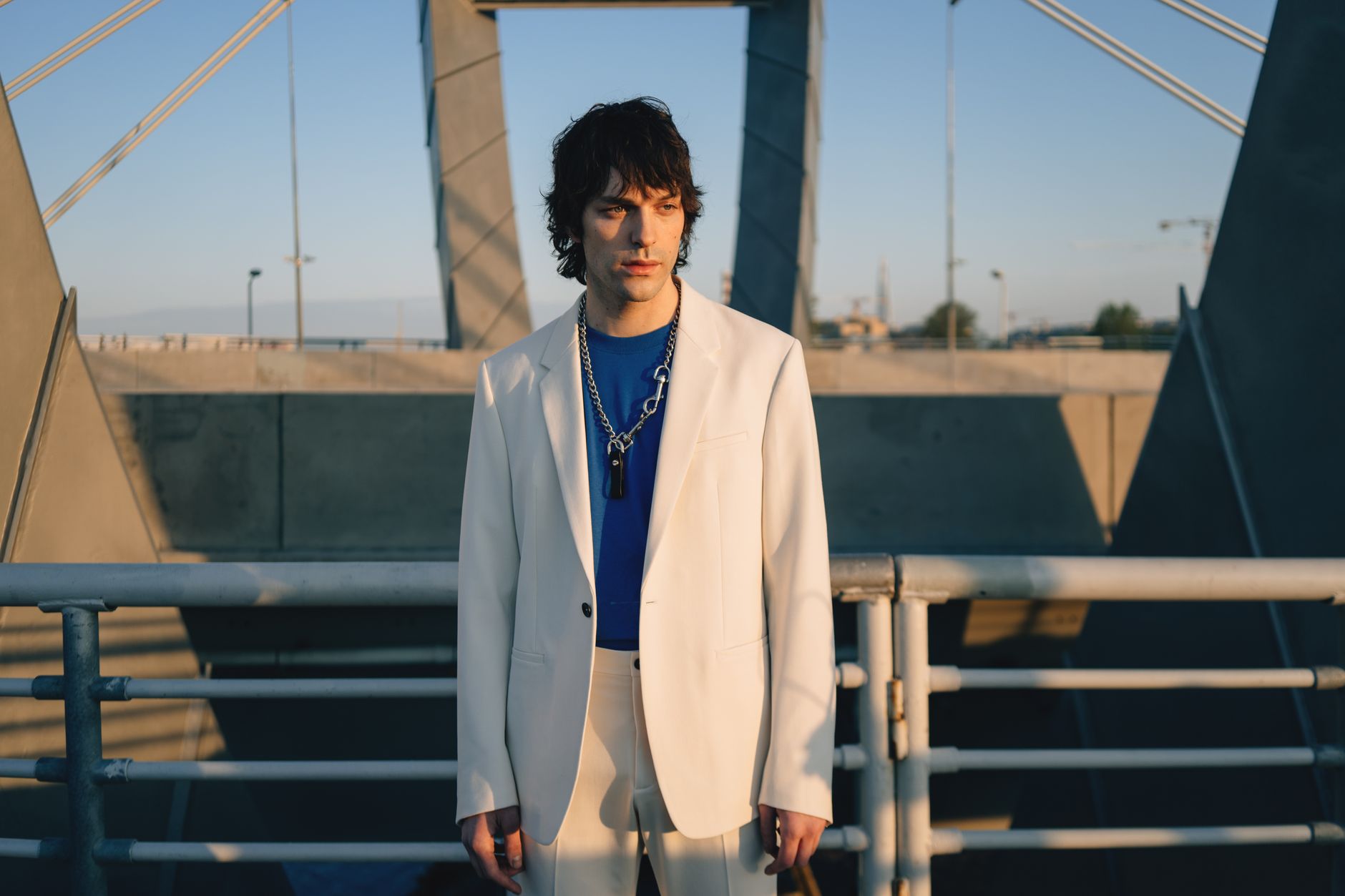 Fashion-forward man in a white blazer poses on a modern bridge under the sun.
