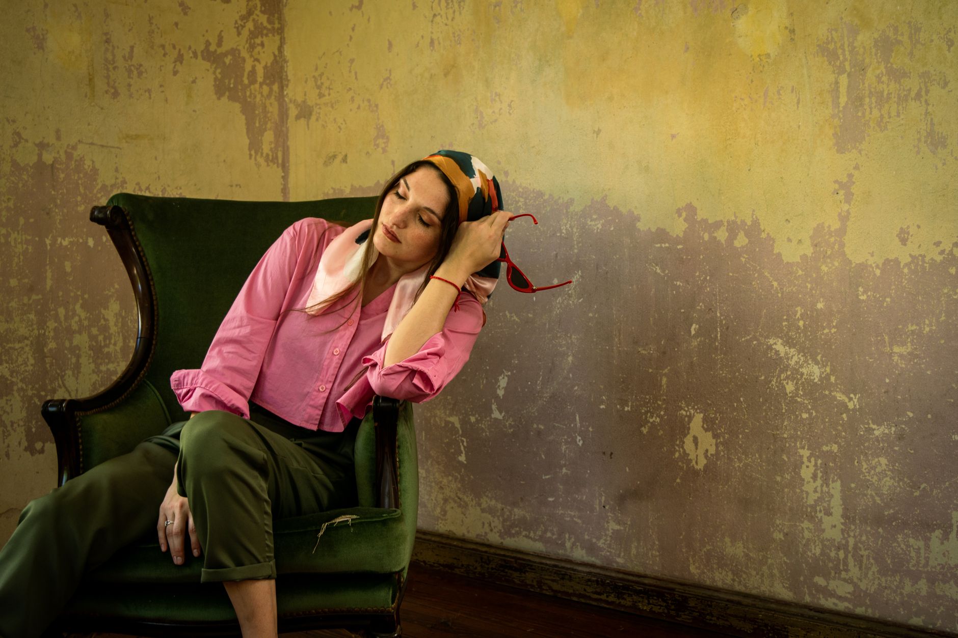 Elegant woman with silk scarf seated on a vintage green chair in a rustic room.