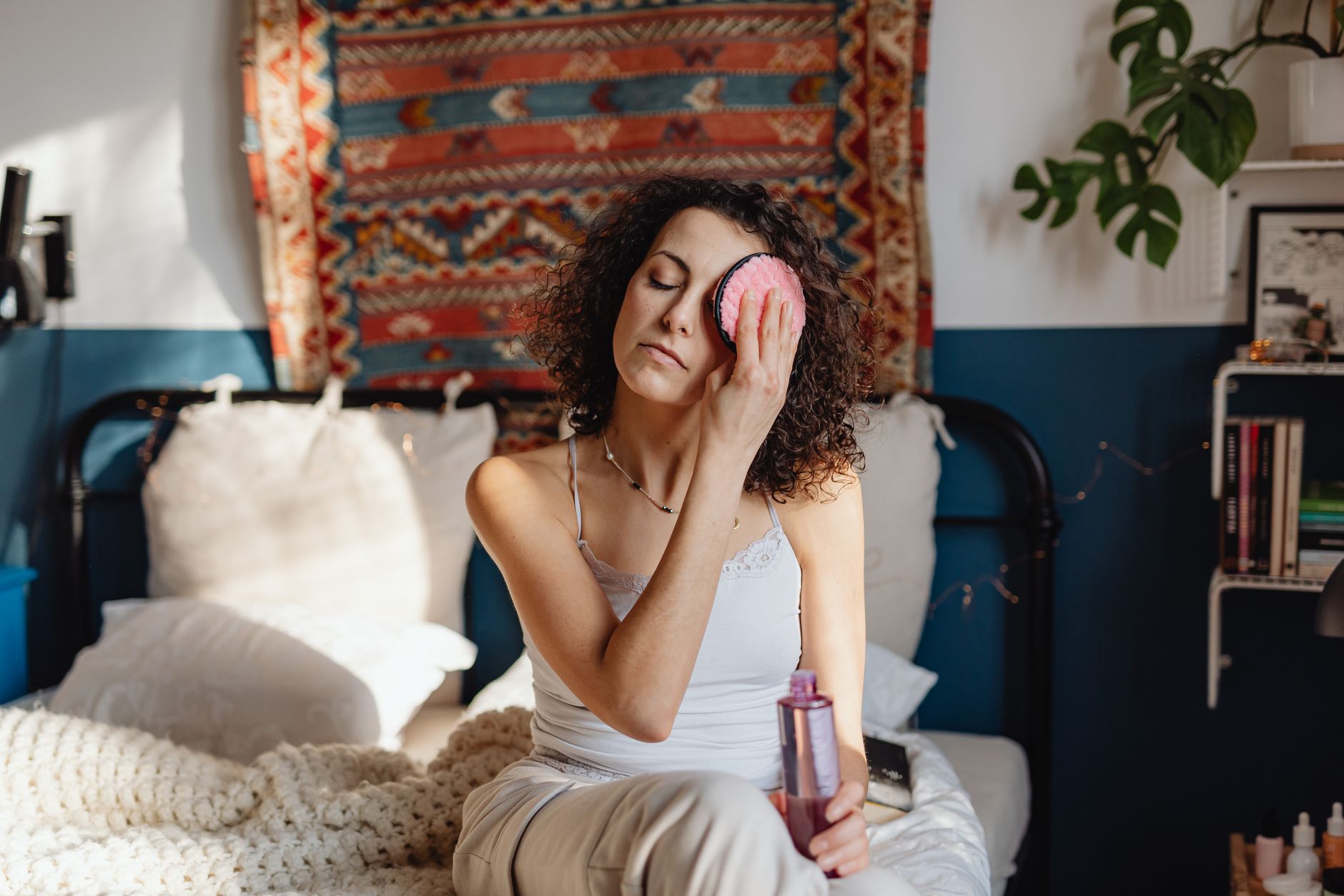 Woman with curly hair removes makeup using a cotton pad in a cozy, stylish bedroom.