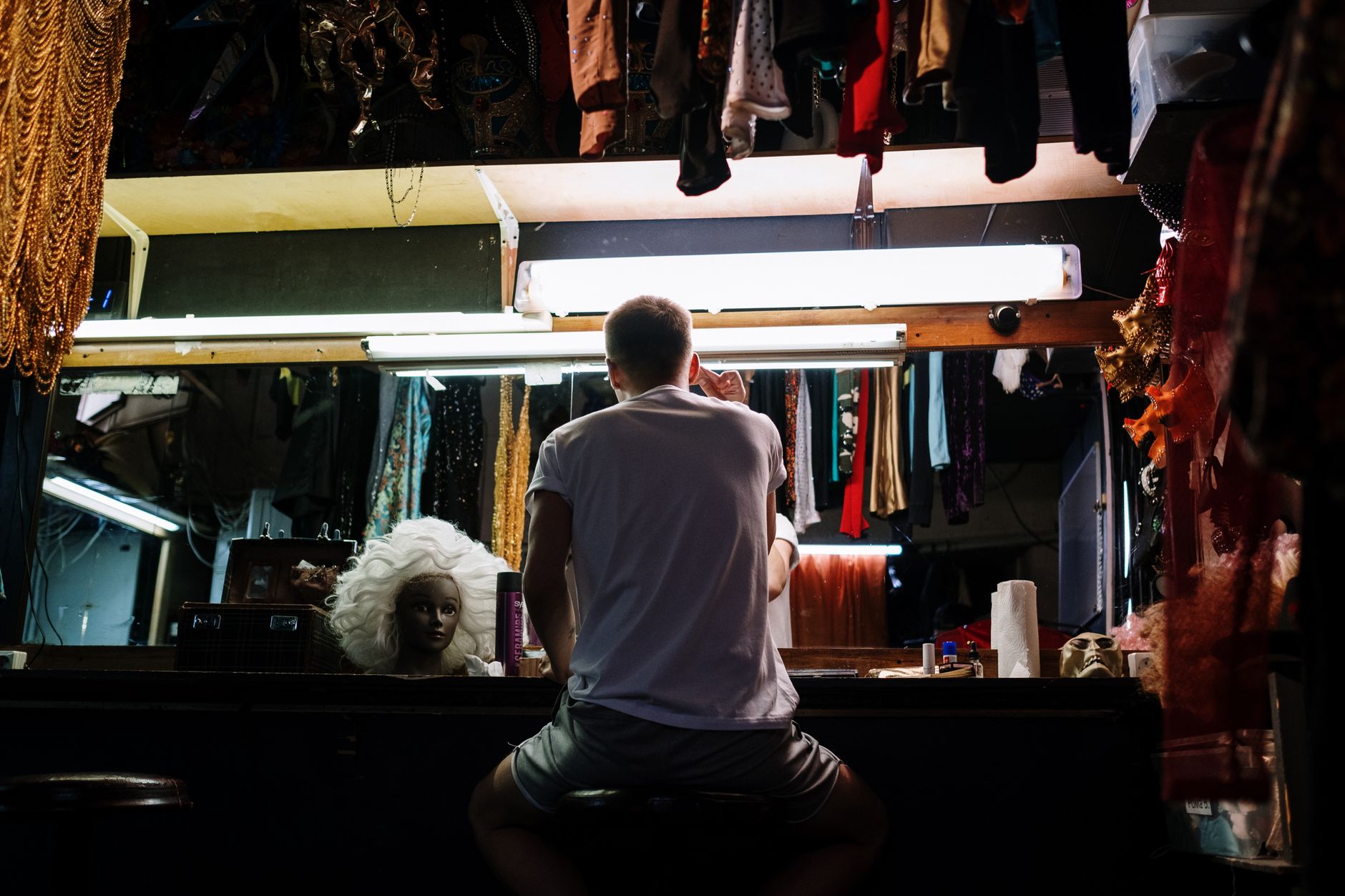 An anonymous person preparing backstage with costumes, wigs, and mirrors in a dressing room.