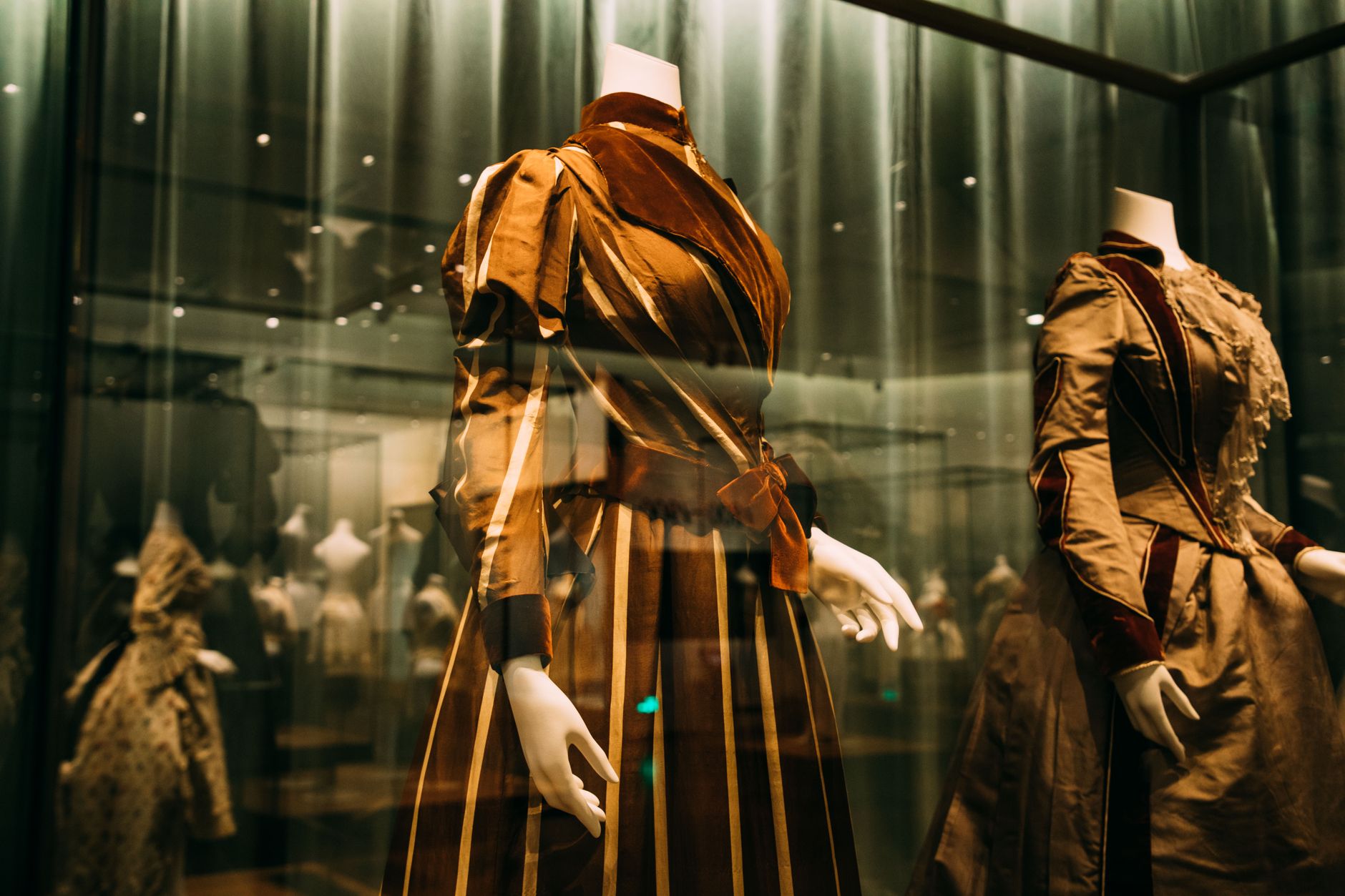 Through glass view of bright ornamental aged style feminine dresses on mannequins in museum