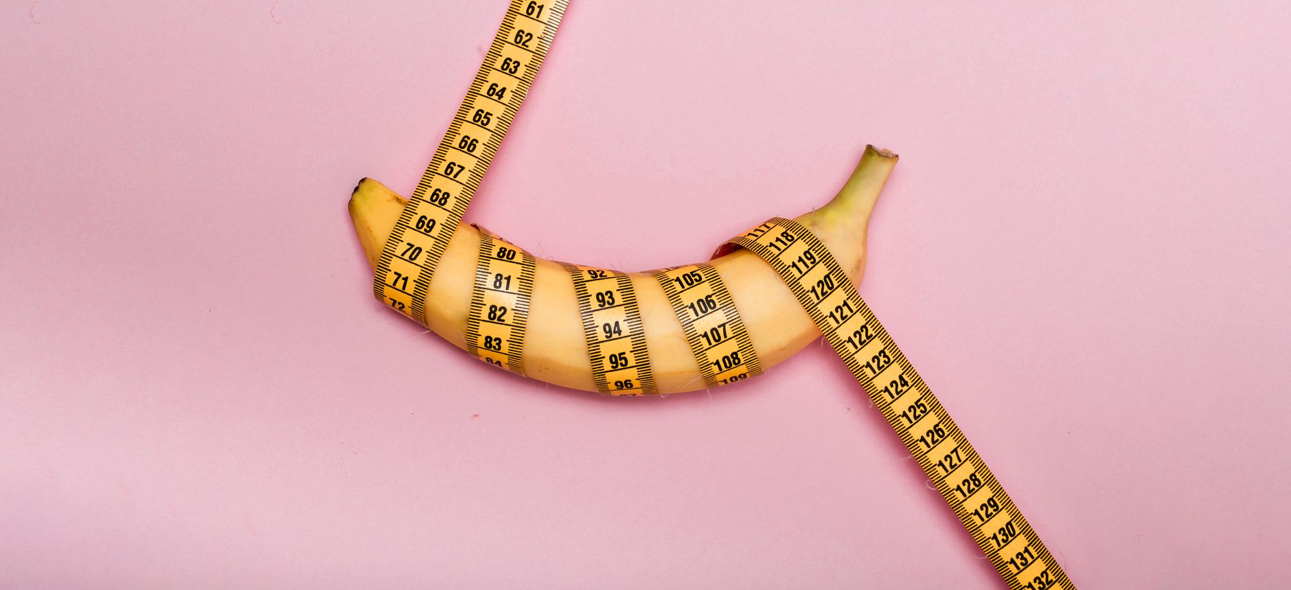A banana wrapped with measuring tape on a pink background representing measurement or size concept.