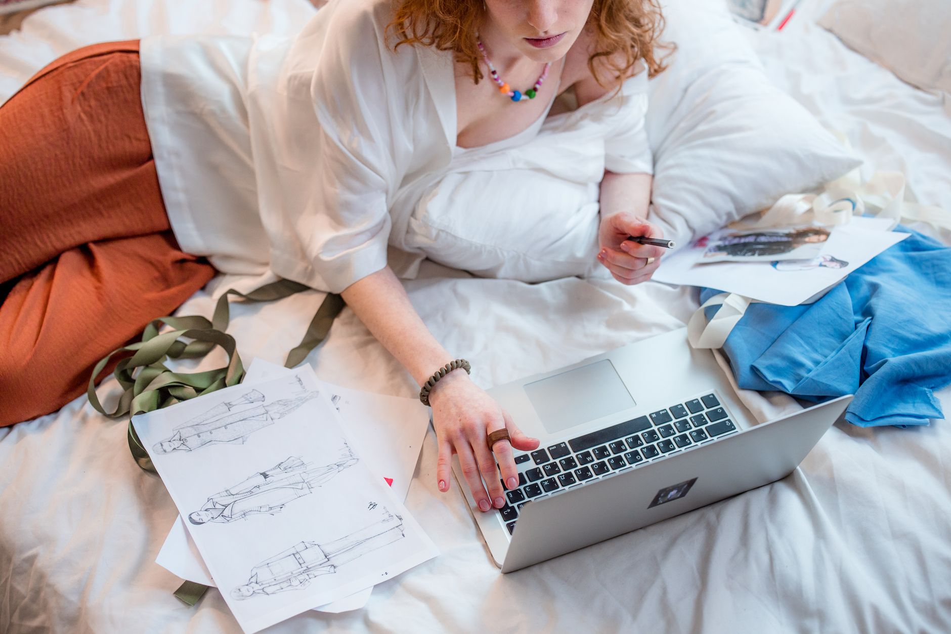 Fashion designer lying on bed with laptop and sketches, working remotely in a cozy home setting.