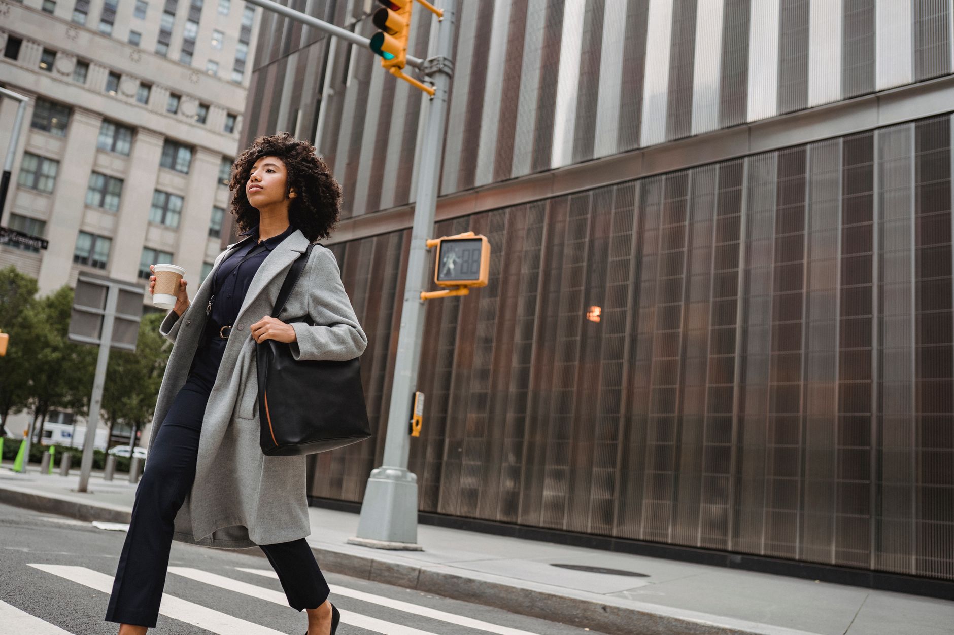 Femme en manteau gris traversant une rue, un grand sac noir à l’épaule et un gobelet de café à la main, devant des immeubles.