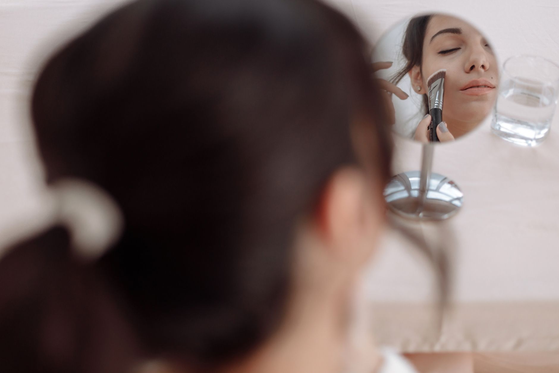 Femme vue de dos se maquillant avec un pinceau devant un petit miroir rond, son visage reflété les yeux fermés, un verre d’eau à côté.
