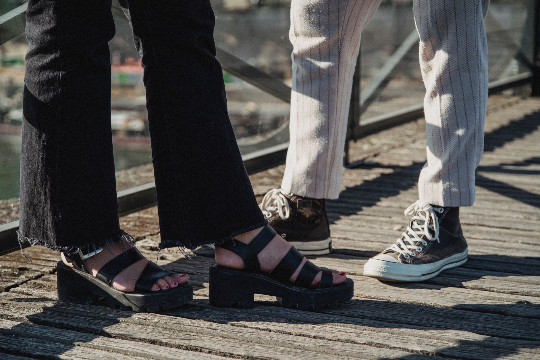 Pieds de deux personnes sur une passerelle en bois : sandales noires à talon épais à gauche, baskets montantes sombres à droite.